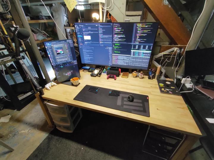 A butcher-block desk in a basement under the stairs with a very large monitor full of code in the middle, a smaller monitor turned vertical to its left, a split keyboard and mouse on a desk mat, and a microphone on a boom arm