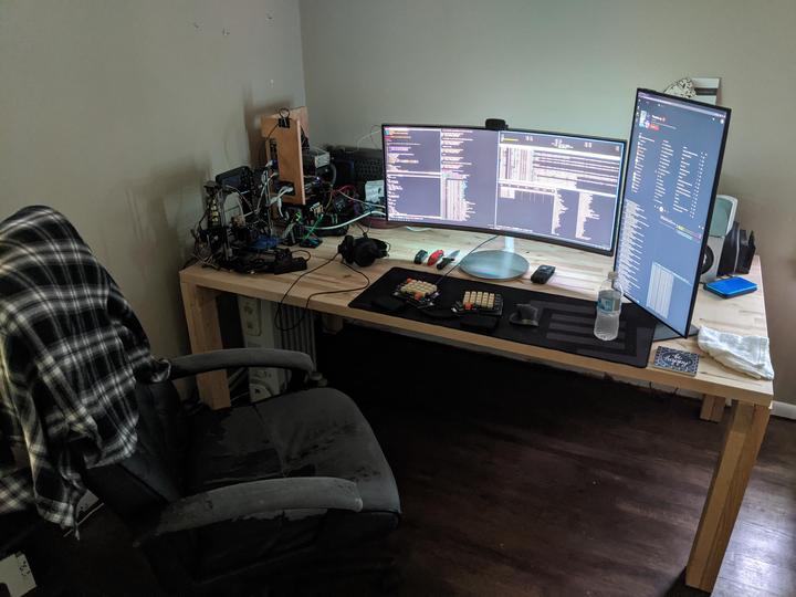 A long, unfinished wooden desk in a spare room holding a curved ultrawide monitor, a second monitor turned vertical, a split keyboard on a desk mat, and a tangle of electronics projects at one end