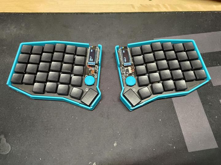 The two halves of a split keyboard with black low-profile keycaps in bright blue cases, each half with a small display and a rotary knob near the thumb keys